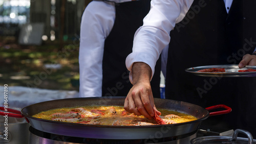 Chef preparing paella outdoors, adding langoustine to large pan
