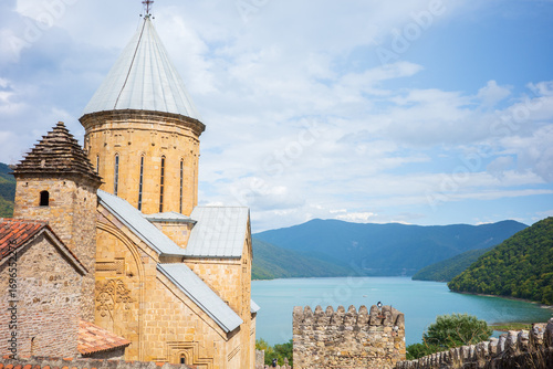 A scenic view of a historic stone church in Georgia with a tall dome roof, ancient fortress walls, and stunning turquoise lake surrounded by lush green mountains. A perfect medieval architecture