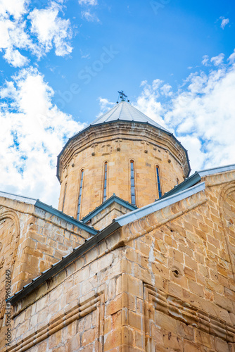 A stunning view of an old Georgian Orthodox church dome made of golden stone, rising gracefully beneath a bright blue sky with scattered clouds. Perfect architectural travel photography in Georgia
