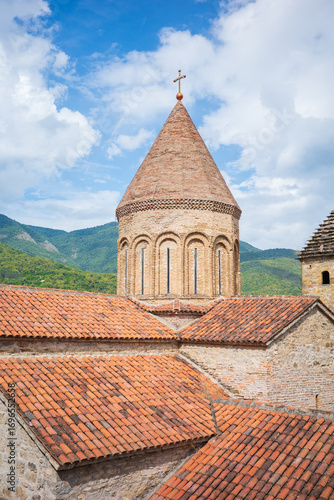 A beautiful view of a historic Georgian church with a conical brick dome topped by a cross, surrounded by red tiled rooftops and lush green mountains. A perfect blend of medieval architecture, culture