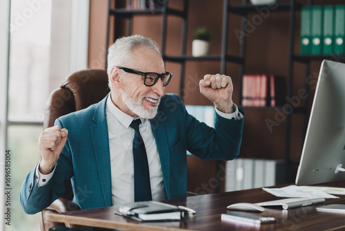 Mature businessman in formal suit celebrating success while working in a corporate office environment with a computer.