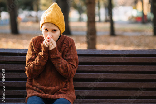 The boy sitting on bench in park in cold autumn day. Kid is warming his hands.