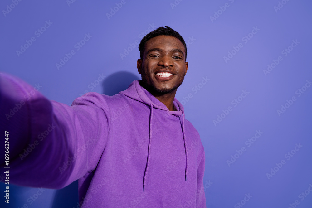 Fototapeta premium Portrait of a smiling young man in a purple hoodie posing against a vibrant violet background, showcasing casual style