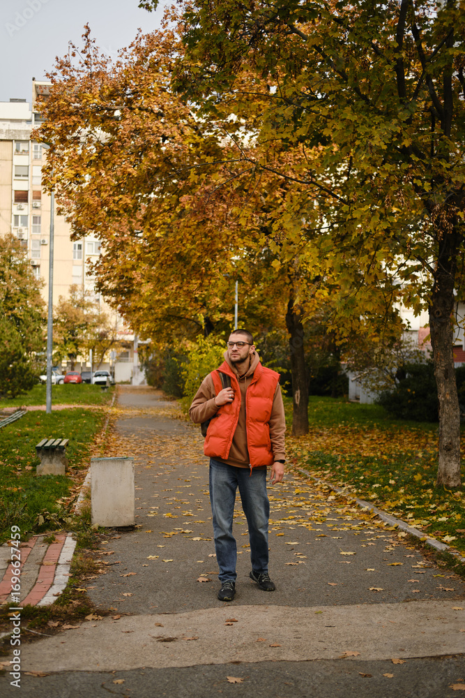 Obraz premium Man in orange vest walking along a tree-lined path covered with autumn leaves
