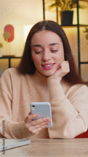 Caucasian young woman sits at table uses mobile phone smiles at home office room apartment. Girl lady texting share messages content on smartphone social media applications online watching relax movie