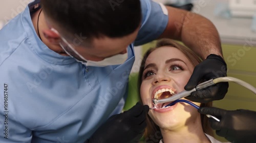 Dentist assistant, Dental equipment, Dental health. During dental treatment, smiling patient is attended by dentist and assistant using multiple tools.