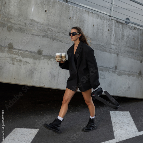 Fashionable young woman in oversized black blazer and sunglasses holding takeaway coffee cups, walking outdoors with confidence and modern street style. Beautiful urban lady with coffee near concrete