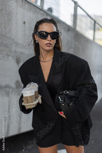Stylish young woman in oversized black blazer and sunglasses walking outdoors with takeaway coffee cups, showcasing modern street fashion and urban lifestyle.