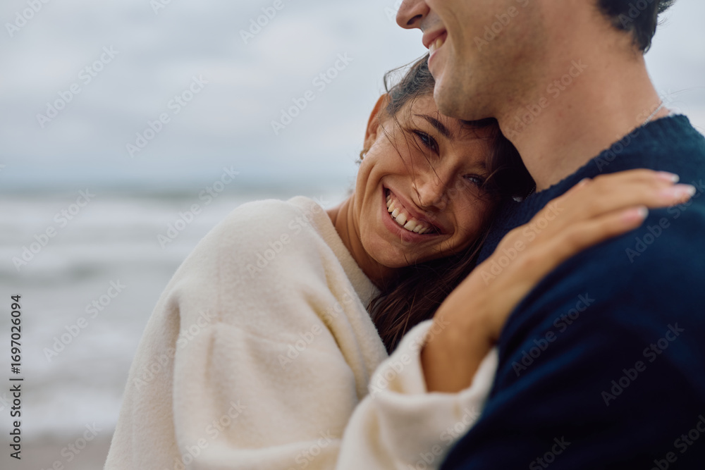 Obraz premium Young woman resting her head on her boyfriend's shoulder, smiling and hugging him on a beach with the sea in the background