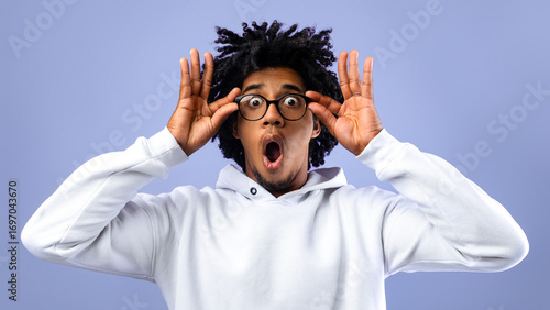 Photography Emotional black teenager opening mouth in shock, touching eyeglasses, looking at camera on blue studio background