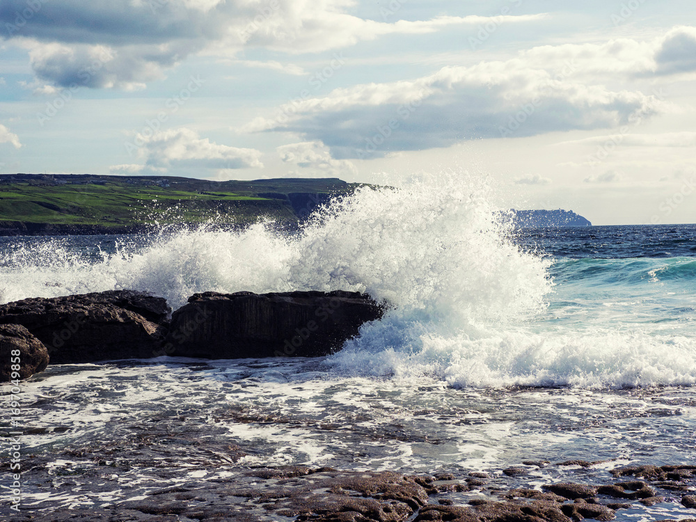 Fototapeta premium Ragged and tough stone coast hit by power full ocean waves, cliff in the background and cloudy sky. Doolin area, county Clare. Ireland. Popular tourist sightseeing area. Irish nature landscape.
