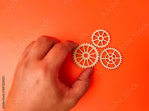The image shows a hand holding a wooden gear, with two other wooden gears lying on an orange background