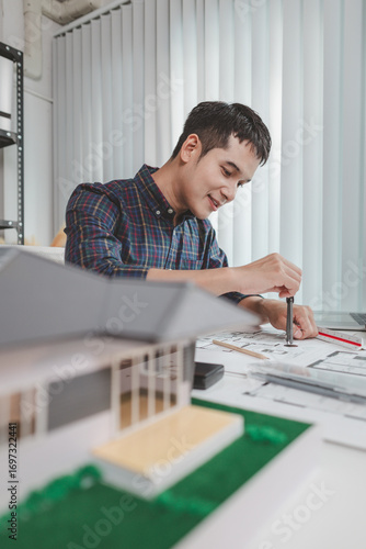 Young male architect working at office desk, drawing construction plans and using drafting tools, focused on building design while showing creativity and dedication in architecture.