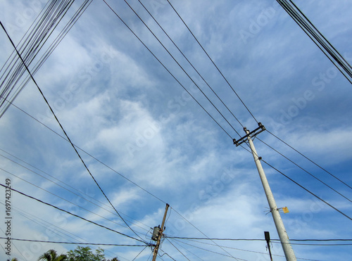 Wallpaper Mural A complex web of crisscrossing electrical wires and cables stretches between two utility poles, set against a calm blue sky with soft, diffuse clouds, showing a typical urban landscape view. Torontodigital.ca