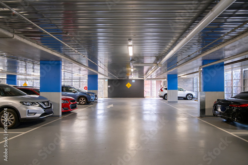 Indoor covered car park with parked vehicles and structural columns within a multi-level facility. Modern urban parking infrastructure with ventilation