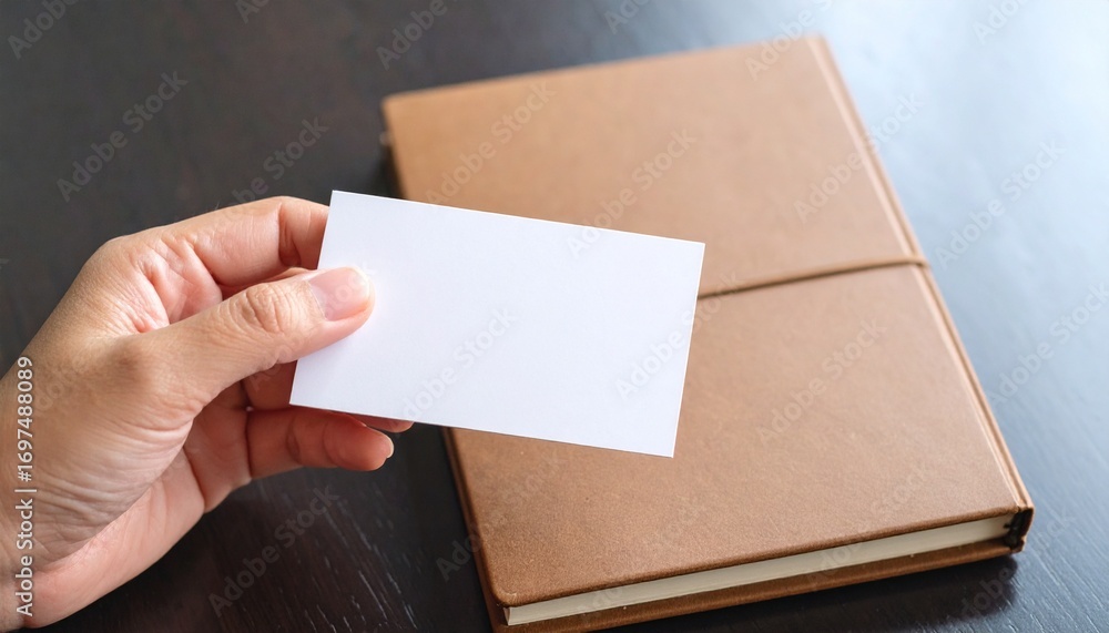 Obraz premium A person's hand holding a blank white business card over a brown journal, ideal for a professional branding mockup
