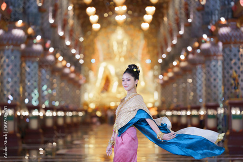 Elegant Thai pretty woman in traditional costume posing gracefully inside a golden temple hall.