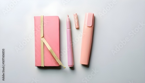 A pink notebook with a gold ribbon, a matching pink pen, and a peach-colored highlighter, all arranged neatly on a clean white background. Perfect for study, office, or creative wor