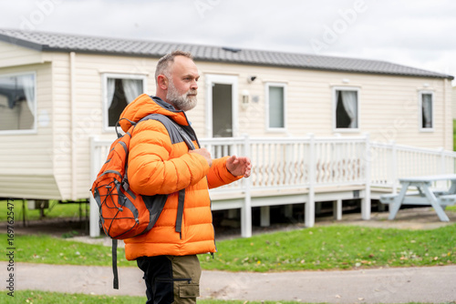 Middle-aged man in bright orange jacket and backpack walking near a mobile home at a campsite during an overcast day