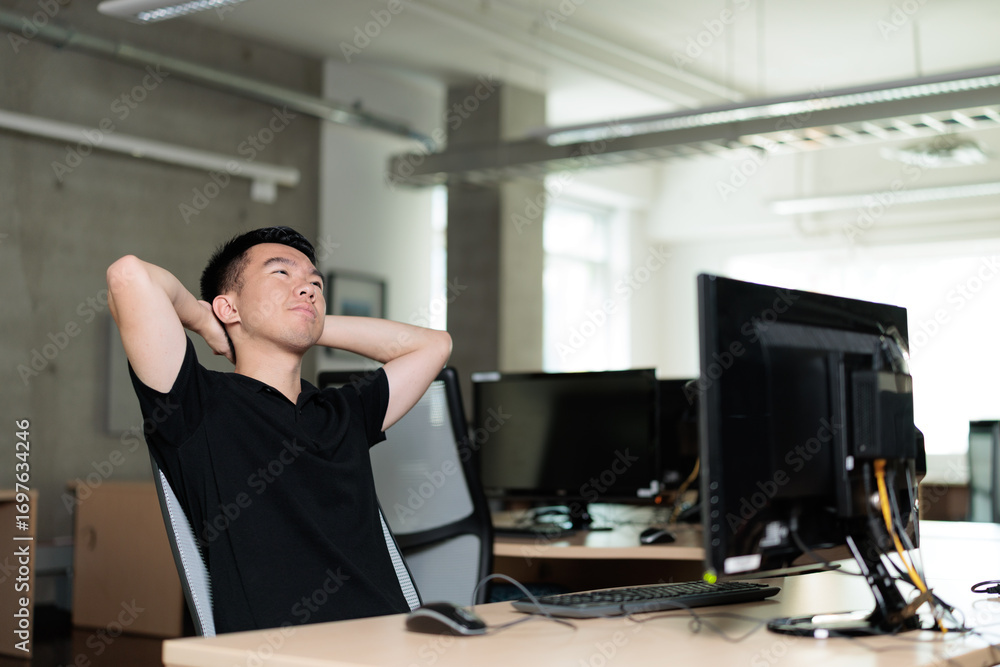 Fototapeta premium A young man leans back in his chair with hands behind his head, gazing upward in thought. He is daydreaming in the office.