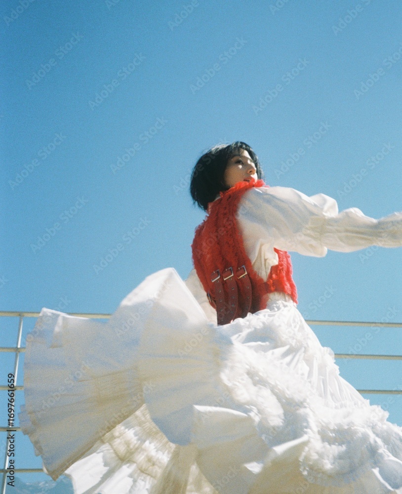 Φωτογραφία A woman in a red vest and white flowing dress twirls against a blue sky backdrop