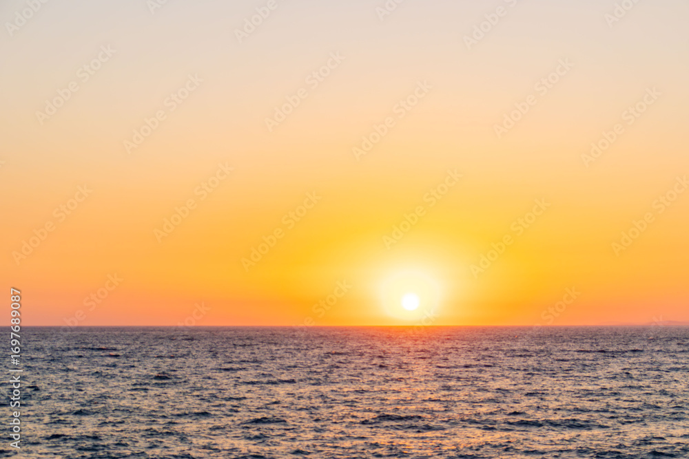 Fototapeta premium Beautiful view of a summer sunset over the sea horizon. The glowing sun creates golden reflections on the water, filling the sky with warm orange and yellow tones. Symbol of calmness, romance