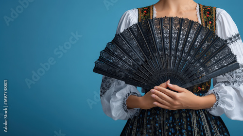 Traditional folk costume with embroidered blouse and decorative black lace fan held by woman on blue background