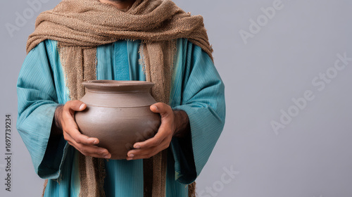 Traditional clay pot held by person wearing blue robe and brown scarf, close up of hands showing cultural craftsmanship and heritage