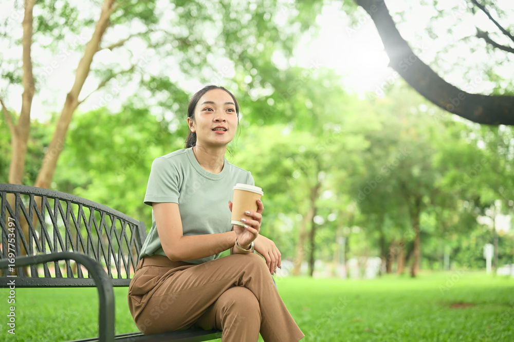 Obraz premium Young woman sitting on a park bench with a takeaway coffee cup, enjoying a peaceful outdoor moment