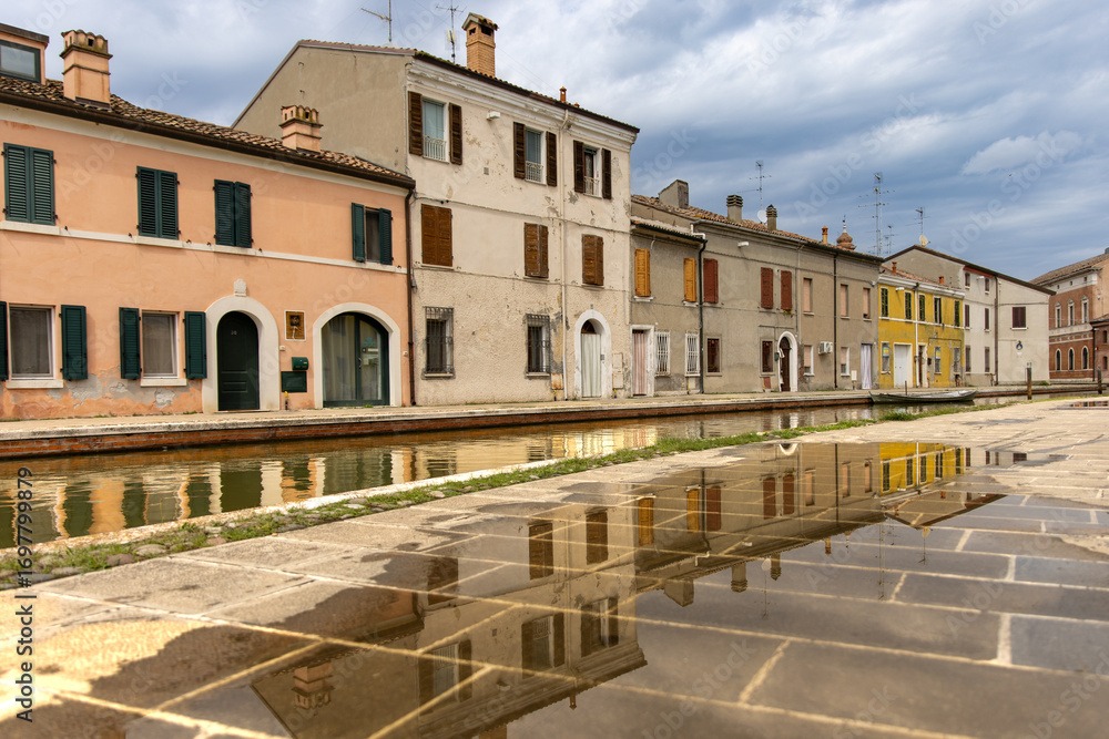 Obraz premium Comacchio, Italy 08 24 2025 colorful canal houses reflected in puddles and water, showcasing pastel facades, shutters and historic charm of Emilia Romagna’s old town under dramatic cloudy sky.