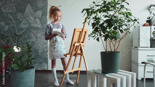 Little girl painting on easel with bright palette and focused face. Concept of early creativity, artistic expression, and talent development in childhood. Indoor natural scene