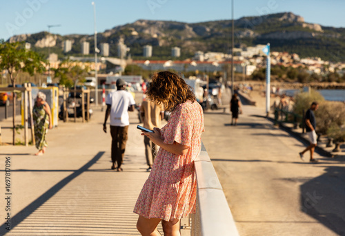 Jeune femme tapant sur son smartphone, vue de profil sur un trottoir le long de la mer, portant une robe d'été fleurie, adossée à une rambarde, promeneurs, ville et colline en arrière-plan, Marseille