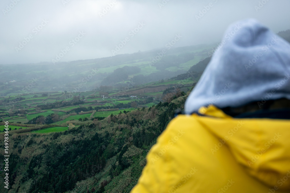 Naklejka premium A person in a yellow jacket looking at a foggy landscape in the Azores The Spirit of Contemplation