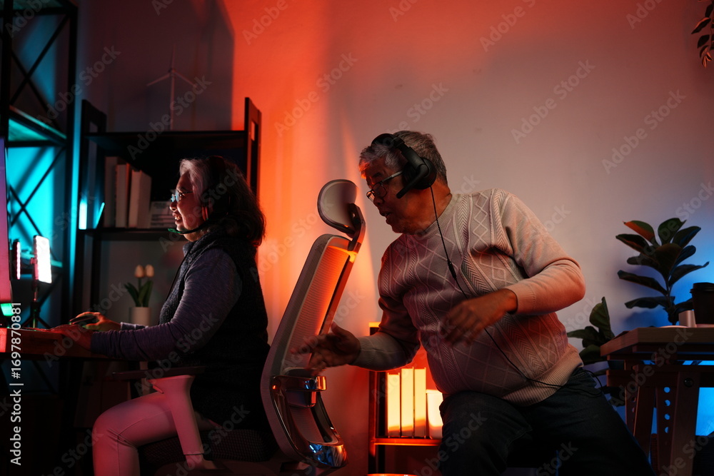 Fototapeta premium Elderly people have a hobby of playing esports. An Asian elderly woman and her elderly husband happily win online games, look at the screen, and excitedly go to the game room with colorful lights.
