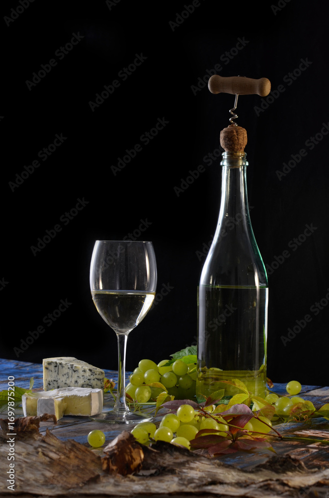 Obraz premium Bottle of white wine with corkscrew, glass of white wine, green grapes and blue cheese on black background.