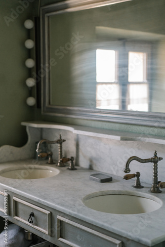 Vintage double sink bathroom vanity with marble countertop, antique brass faucets, and a large wall mirror. Soft natural light creates a calm and rustic atmosphere.