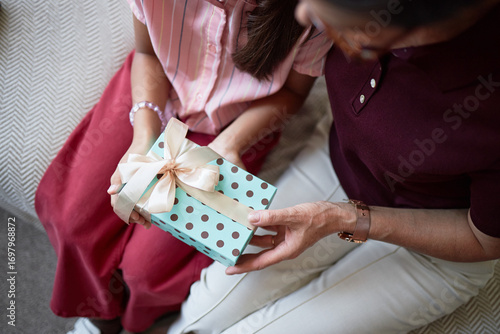 Caucasian middle aged woman giving polka dot gift box with ribbon to Caucasian child girl sitting together on sofa, hands holding present, close up of interaction