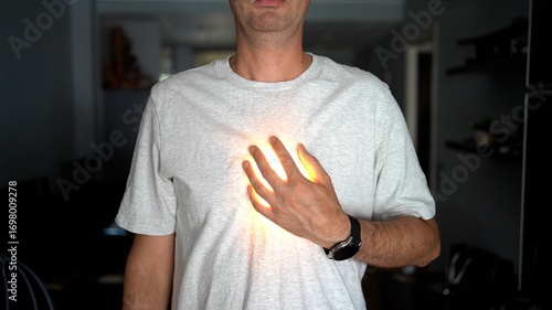 Photos A man in a white t-shirt places his hand on his chest with a glowing light effect under his hand, symbolizing heart, energy, or inner power