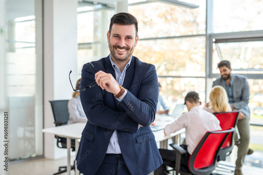 Fototapeta premium Confident businessman with glasses smiling at camera standing in office with colleagues in background