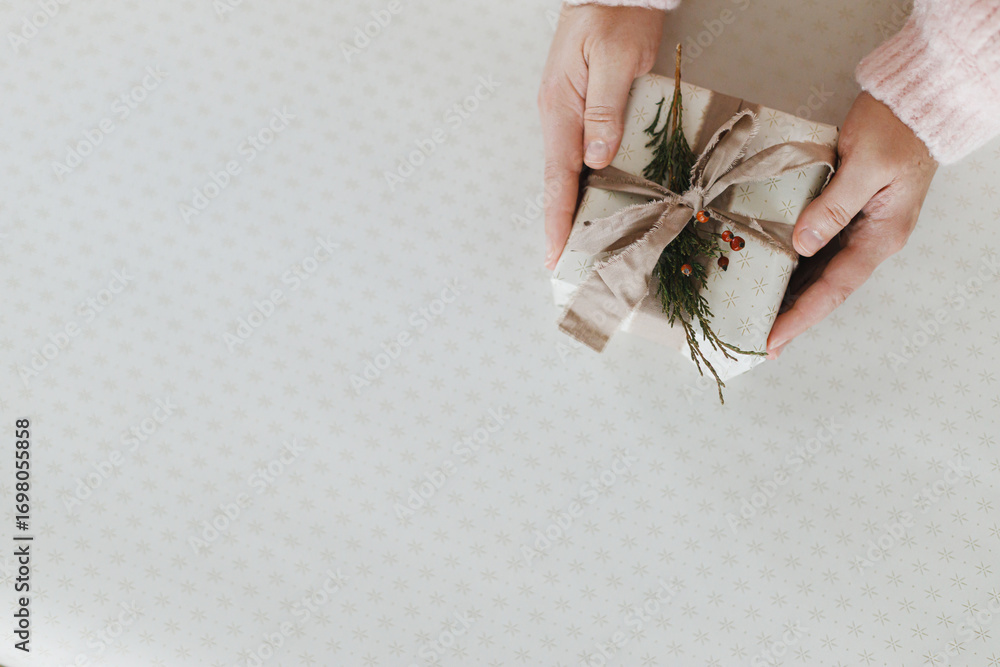 Fototapeta premium Merry Christmas and Happy Holidays! Hands holding stylish christmas present on festive paper flat lay. Xmas gift in with linen ribbon and cedar branch, copy space. Holiday shopping
