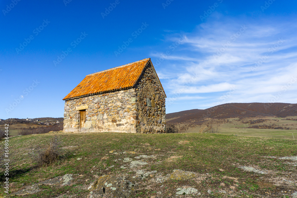 Fototapeta premium Small shurch on the rock. Wooden door. Orange tiled roof, stone walls. Green grass and bushes. Bright blue sly with clouds. Georgia