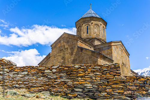 Gergeti Holy Trinity Church. Stone wall fence, dry masonry, orange lichen, stone walls with bas-reliefs, dome with a cross. Bright blue sky with clouds.
