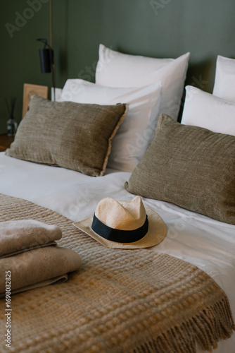 Cozy hotel bedroom with neatly folded towels on the bed, decorative pillows and a straw hat, stylish hospitality concept. Warm neutral tones create a welcoming and relaxing atmosphere.