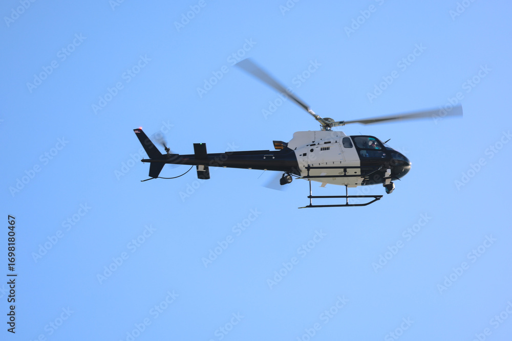 Fototapeta premium Black and white helicopter flying against a clear blue sky, captured in motion with spinning rotor blades.