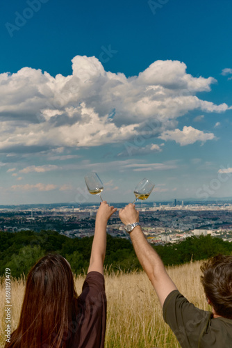 Back view of a couple in tall yellow-green grass, raising glasses of white wine. Romantic picnic moment, sunny day with panoramic view of Vienna, Austria.