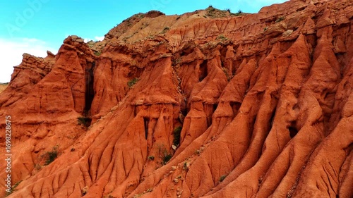 Drone lifts off from the red formations of Skazka Canyon in Kyrgyzstan, slowly rising to unveil a breathtaking mountain ridge in the distance under a bright sky.