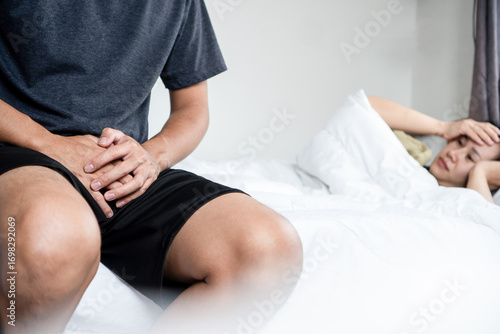 Asian couple in bed struggling with intimacy problems and relationship stress caused by erectile dysfunction.  husband sits on bed holding his crotch and frustrated, the wife looking worried, unhappy