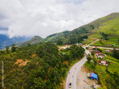 A dramatic aerial view of the mountain landscape in Flesha, Ainaro, Timor-Leste. A winding road passes through a remote village nestled among lush green hills.