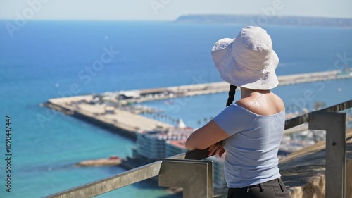 A traveler girl in a white bucket hat takes in the coast from a breezy hilltop lookout in Alicante, Spain