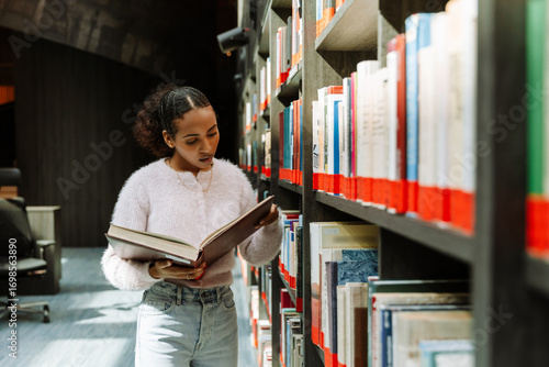 A female student is reading a book that she is holding in her hands while standing by the bookshelves